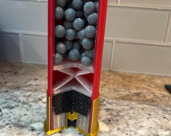 Op de afbeelding: Een doorsnede van een rode en gouden hagelpatroon, die de interne componenten laat zien. De patroon is gevuld met talrijke grijze pellets. De basis van de patroon is goudkleurig en de interne onderdelen zijn wit en zwart.