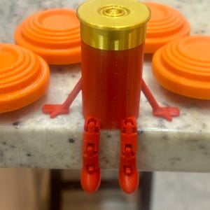 Puede incluir: Un personaje de cartucho de escopeta rojo y dorado impreso en 3D con brazos y piernas, rodeado de platos de arcilla naranja. El cartucho tiene una parte superior dorada y está posado como si estuviera sentado. Los platos de arcilla son circulares y naranjas.