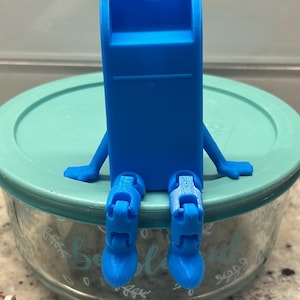 May include: A bright blue, 3D-printed whistle with arms and legs, sitting on a teal-colored lid of a glass container. The whistle has a rectangular body with a rectangular opening at the top. The container has a floral design.