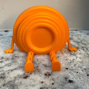 Puede incluir: Objeto naranja impreso en 3D, con cuerpo circular y pequeños brazos y piernas. El objeto está tumbado boca abajo, con las piernas extendidas. El diseño es juguetón y de estilo caricaturesco.