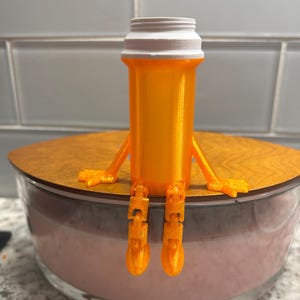 May include: A whimsical orange pill bottle figurine with articulated arms and legs, sitting on a wooden lid atop a clear glass bowl. The bottle has a white cap and is made of a translucent orange material.