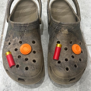 Op de afbeelding: Een paar bruine camouflage Crocs versierd met oranje en rode bedels. De bedels omvatten twee oranje kleiduivendoelen en twee rode hagelpatroonbedels. De schoenen staan op een grijze tegelvloer.