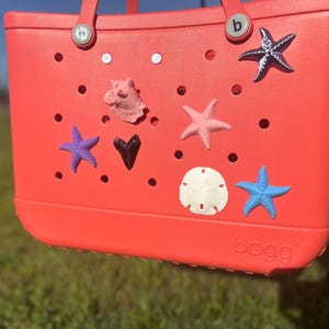 Pode incluir: Uma bolsa tote cor-de-coral decorada com vários encantos com tema marinho. A bolsa apresenta uma coleção de estrelas do mar, uma concha e uma estrela-do-mar. A bolsa tem uma alça e a marca "bogg" é visível.