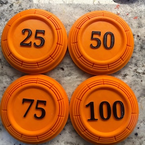 May include: Four orange clay targets with black numbers: 25, 50, 75, and 100. Each target has a circular design with concentric rings. The targets are used for shooting sports.