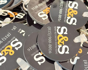May include: A close-up of a pile of black and white round keychains with the text "S&S" and a phone number. The keychains are made of a durable material and have a clear plastic ring for attaching keys.