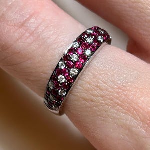 May include: A silver ring with a band covered in alternating rows of small, round, red and white gemstones. The ring is worn on a finger, showcasing the intricate design and sparkle of the gemstones. The gemstones are set closely together, creating a continuous pattern.