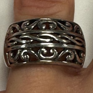 May include: A silver-colored ring with an intricate openwork design. The ring features a series of swirling, scroll-like patterns that create a textured surface. The ring is wide and sits on a finger, showcasing its detailed craftsmanship.