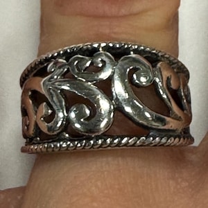 May include: A silver ring with an openwork design featuring swirling, scroll-like patterns. The ring has a textured, rope-like border on the top and bottom edges. The ring is worn on a finger.