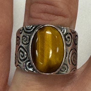 May include: A silver ring with a large, oval tiger's eye stone. The band features a detailed swirl design. The ring is shown on a finger.