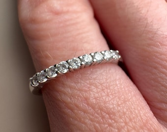 May include: A silver band ring with a row of small, clear gemstones. The ring is worn on a finger, showcasing the sparkling stones. The ring's design is simple and elegant, suitable for everyday wear or special occasions.