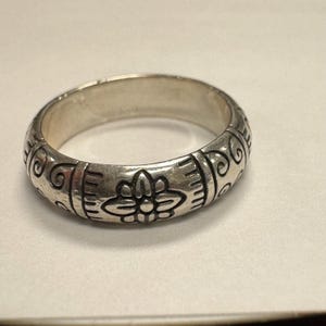 Op de afbeelding: Een zilveren ring met een bloemen- en krulontwerp. De ring heeft een brede band met ge&euml;tste details. Het bloemmotief is gecentreerd, met krul- en lijnpatronen aan weerszijden. De ring is gemaakt van een glanzend, metalen materiaal.