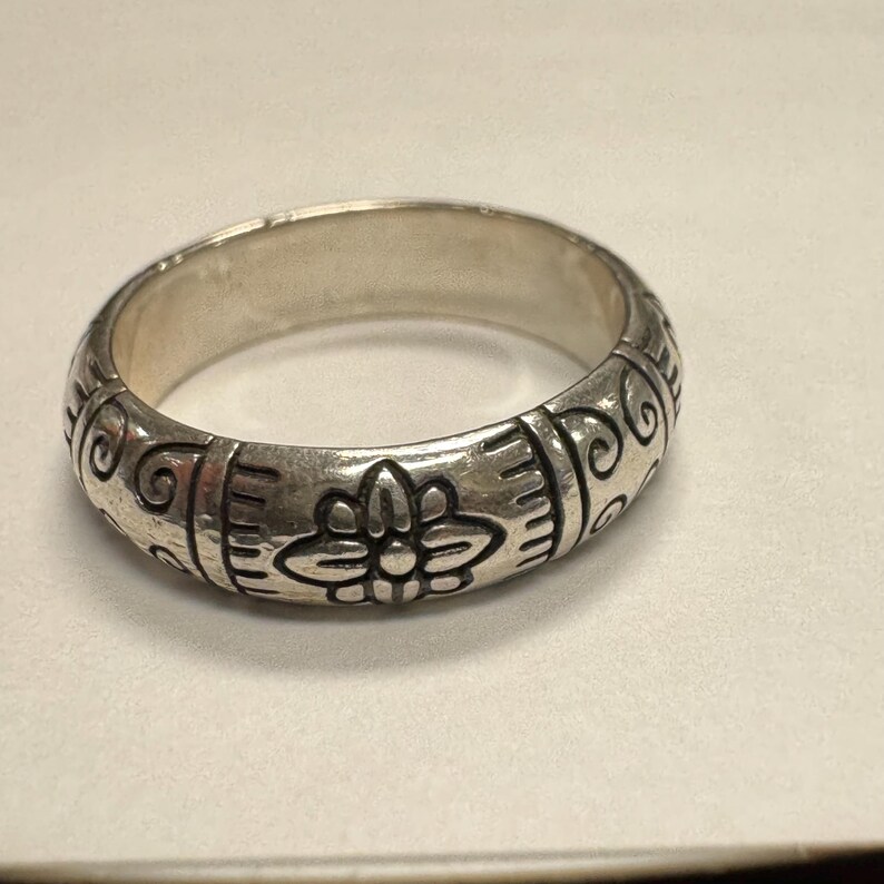 Op de afbeelding: Een zilveren ring met een bloemen- en krulontwerp. De ring heeft een brede band met ge&euml;tste details. Het bloemmotief is gecentreerd, met krul- en lijnpatronen aan weerszijden. De ring is gemaakt van een glanzend, metalen materiaal.