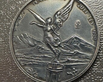 May include: A silver coin featuring a winged figure with outstretched arms, mountains, and the text "PLATA PURA" and "1 ONZA". The coin also has the year "2011" and the fineness "999" inscribed on it. The coin is round and has a detailed design.