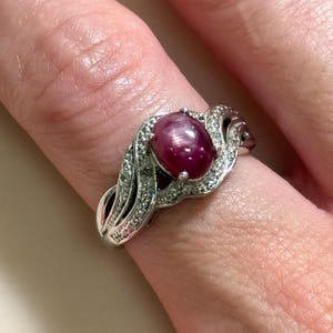 May include: A silver ring featuring a large, oval, deep red gemstone, possibly a ruby, at the center. The gemstone is surrounded by smaller, clear stones. The ring's band has a decorative, swirling design.