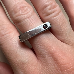 May include: A silver-toned metal ring featuring a flat, rectangular bar design across the top. The surface of the bar is smooth and reflective, accented by a single, small, round, deep red gemstone set near one end of the rectangular piece.