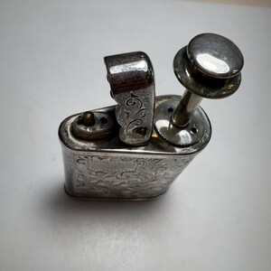 May include: A silver-toned metal table lighter featuring an intricate engraved floral pattern across its body. The lighter has a vertical plunger mechanism on top, a hinged lid, and a small circular ignition nozzle on the flat upper surface.