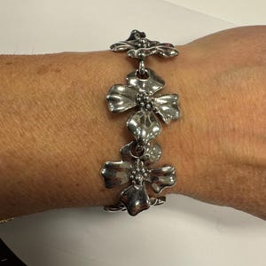 May include: A silver-toned bracelet featuring a floral design. The bracelet is composed of multiple flower-shaped links connected by small rings. The flowers have detailed petals and a textured center, creating a three-dimensional effect. The bracelet is worn on a wrist.