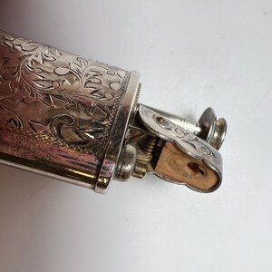 May include: A silver-toned metal lighter featuring an intricate, engraved floral pattern across its cylindrical body. The mechanism includes a hinged top with matching engravings and a visible wooden insert at the ignition point, showing a metallic finish.