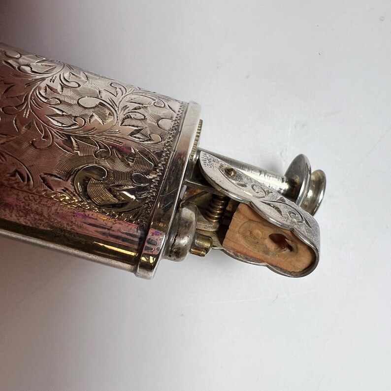 May include: A silver-toned metal lighter featuring an intricate, engraved floral pattern across its cylindrical body. The mechanism includes a hinged top with matching engravings and a visible wooden insert at the ignition point, showing a metallic finish.