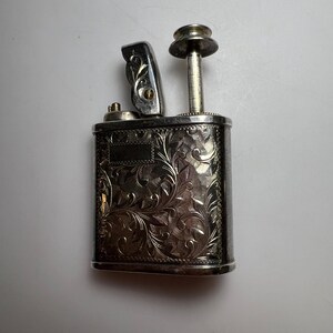 May include: A silver-toned metal table lighter featuring an intricate engraved floral pattern across its rectangular body. The lighter includes a vertical ignition lever and a tall, slender cylindrical fuel cap mechanism extending from the top surface.