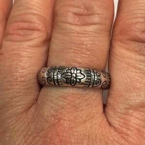 Op de afbeelding: Een zilveren ring met een bloemenpatroon. De ring heeft een centraal bloemmotief, geflankeerd door verticale lijnen en krullen. De ring wordt aan een vinger gedragen.