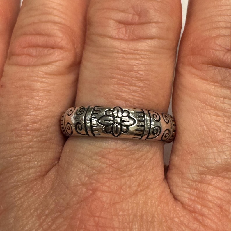 Op de afbeelding: Een zilveren ring met een bloemenpatroon. De ring heeft een centraal bloemmotief, geflankeerd door verticale lijnen en krullen. De ring wordt aan een vinger gedragen.