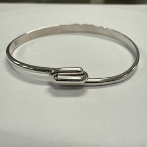 May include: A silver-colored bangle bracelet with a hook closure. The bracelet is a simple, elegant design, with a smooth, polished finish. The hook closure is a unique detail, adding visual interest to the piece.