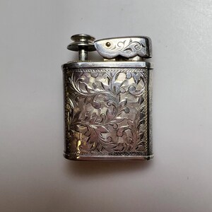 May include: A rectangular silver-toned metal lighter featuring an intricate, engraved floral scrollwork pattern across its front surface. The lighter includes a top-mounted ignition mechanism with a small circular button and a hinged flip-top lid.