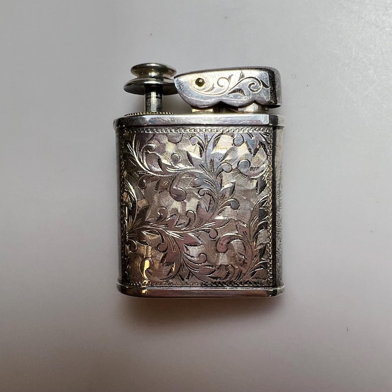May include: A rectangular silver-toned metal lighter featuring an intricate, engraved floral scrollwork pattern across its front surface. The lighter includes a top-mounted ignition mechanism with a small circular button and a hinged flip-top lid.