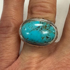 May include: A silver ring featuring a large, oval turquoise stone. The turquoise has a vibrant blue-green color with dark brown matrix patterns. The ring's band has a textured design, adding detail to the jewelry.