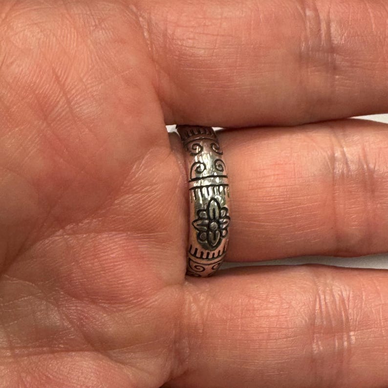 Op de afbeelding: Een zilveren ring met een gegraveerd bloemmotief. De ring heeft een herhalend patroon van krullen en lijnen langs de band. Het centrale ontwerp is een gedetailleerde bloem. De ring wordt aan een vinger gedragen.