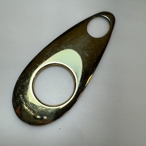 May include: A gold-toned metal bottle opener with a teardrop shape and two circular openings. The opener is marked "GUCCI - MADE IN ITALY". The surface shows minor wear.