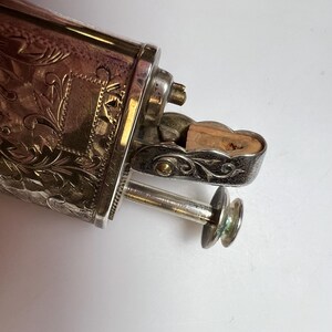 May include: A silver-toned metal lighter featuring an intricate engraved floral pattern across its rectangular body. The mechanism includes a hinged lid with a visible wooden insert and a protruding circular ignition button on a thin metal stem.