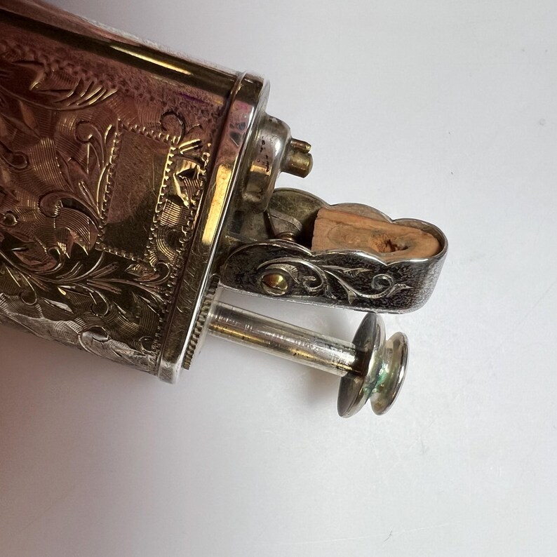 May include: A silver-toned metal lighter featuring an intricate engraved floral pattern across its rectangular body. The mechanism includes a hinged lid with a visible wooden insert and a protruding circular ignition button on a thin metal stem.