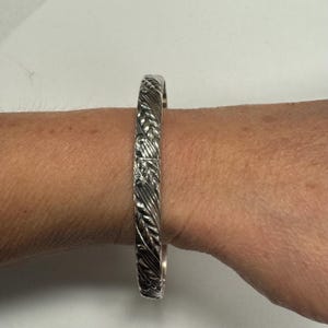 May include: A silver-colored bangle bracelet with a detailed leaf pattern. The bracelet is worn on a wrist, showcasing the intricate design. The metal has a polished finish, reflecting light and highlighting the embossed leaves.