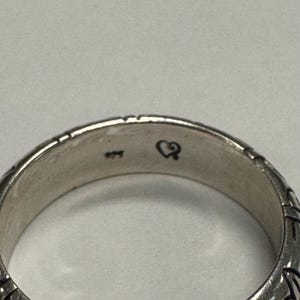 Op de afbeelding: Close-up van een zilveren ring met een textuurontwerp. De ring heeft een klein hartsymbool en het nummer "925" aan de binnenkant gestempeld. Het oppervlak van de ring heeft een gedetailleerd patroon.
