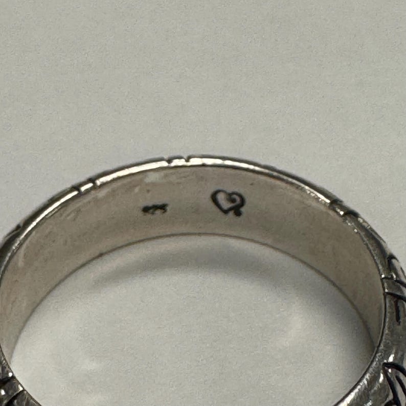 Op de afbeelding: Close-up van een zilveren ring met een textuurontwerp. De ring heeft een klein hartsymbool en het nummer "925" aan de binnenkant gestempeld. Het oppervlak van de ring heeft een gedetailleerd patroon.