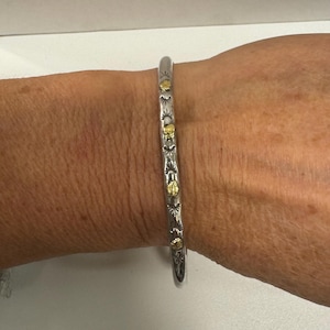 May include: A silver-toned cuff bracelet with gold-colored accents. The bracelet features a series of decorative elements, including sunbursts, butterflies, and small circular shapes. The design is symmetrical and runs along the length of the bracelet.