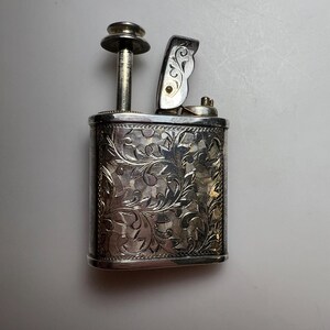 May include: A rectangular silver-toned metal lighter featuring an intricate engraved floral pattern across its front surface. The lighter includes a vertical plunger mechanism on the left side and a hinged lid on the right, both in metallic silver.