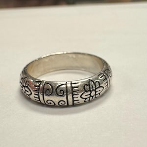 Op de afbeelding: Een zilveren ring met een gegraveerd bloemen- en krulontwerp. De ring heeft een afgeronde band en is waarschijnlijk gemaakt van sterling zilver. De gegraveerde details zijn donkerder gemaakt om het ontwerp te accentueren.