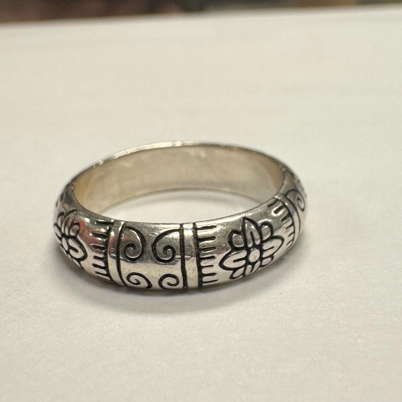 Op de afbeelding: Een zilveren ring met een gegraveerd bloemen- en krulontwerp. De ring heeft een afgeronde band en is waarschijnlijk gemaakt van sterling zilver. De gegraveerde details zijn donkerder gemaakt om het ontwerp te accentueren.
