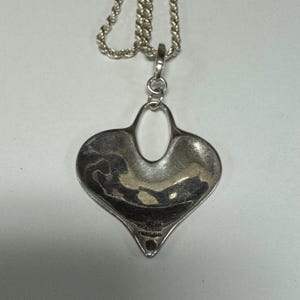 May include: A silver-toned heart-shaped pendant necklace with a textured surface. The pendant has an open oval design in the center and hangs from a silver rope chain. The pendant is marked "925 STERLING THAILAND".