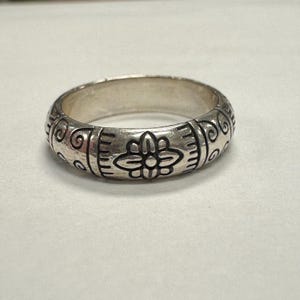 Op de afbeelding: Een zilveren ring met een gegraveerd bloemmotief. De ring heeft een herhalend patroon van gestileerde bloemen en krullen rond de band. De ring wordt getoond op een wit oppervlak.