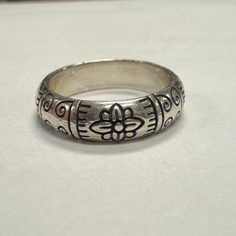 Op de afbeelding: Een zilveren ring met een gegraveerd bloemmotief. De ring heeft een herhalend patroon van gestileerde bloemen en krullen rond de band. De ring wordt getoond op een wit oppervlak.