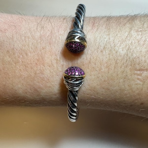 May include: A silver-toned cuff bracelet with a twisted cable design. The bracelet features two decorative ends with a gold band and a cluster of small, round, pink gemstones. The bracelet is shown on a person's wrist.