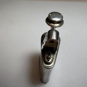 May include: A silver-toned metal lighter with an elongated, rounded rectangular body and a prominent circular push-button mechanism at the top. The lighter features a hinged lid that is currently open, revealing a small brass-coloured nozzle and valve.