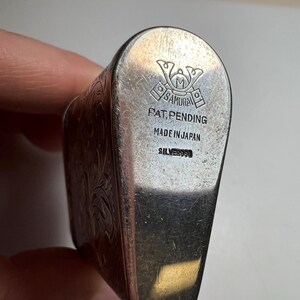 May include: A silver object with an engraved floral pattern on the side. The base features a stamped logo of a helmet with the text SAMURAI, PAT. PENDING, MADE IN JAPAN, and SILVER950. The metal surface shows light scratches and a polished finish.