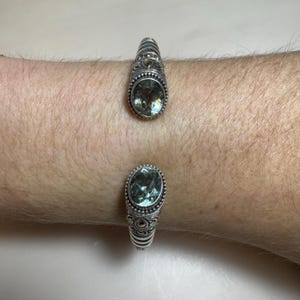 May include: Silver cuff bracelet featuring two oval, light green gemstones. The bracelet has intricate metalwork with a textured design around the gemstones. The bracelet is open, allowing it to be easily put on and taken off.