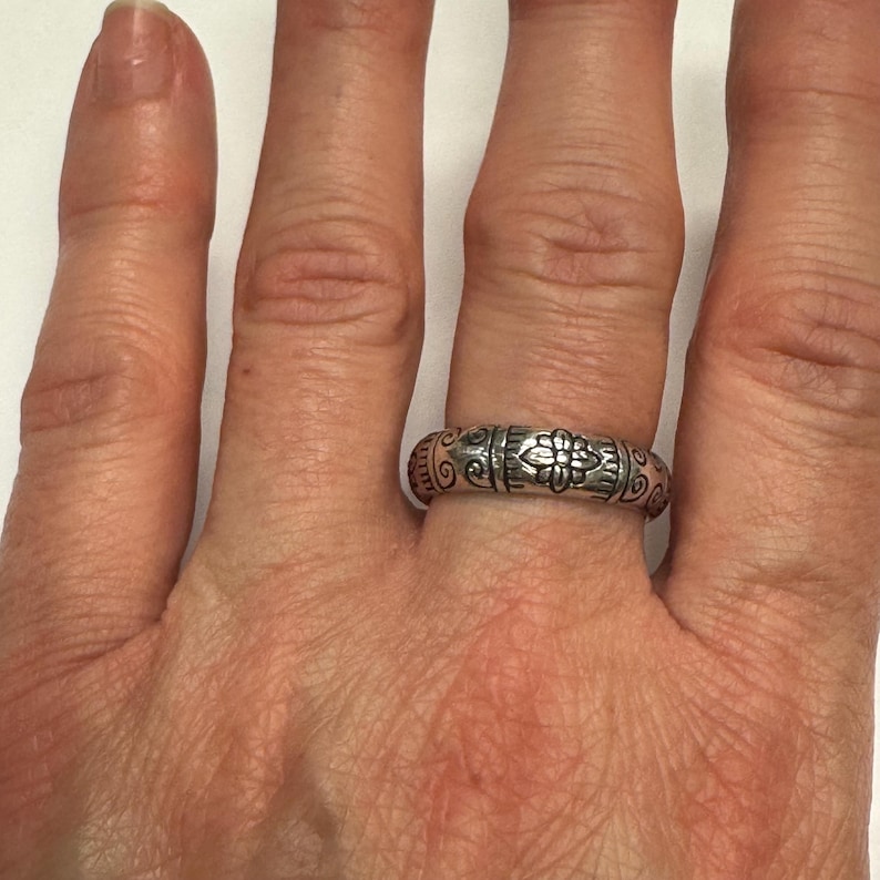Op de afbeelding: Een zilveren ring met een sierlijk bloemen- en krulontwerp. De ring wordt aan een vinger gedragen en heeft gedetailleerde patronen rond de band. Het centrale ontwerpelement is een bloem, geflankeerd door symmetrisch krulwerk.