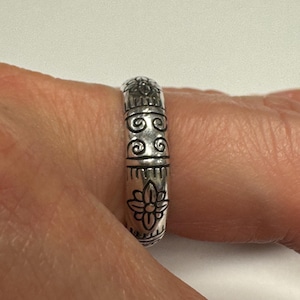 Op de afbeelding: Een zilveren ring met een gegraveerd bloemen- en scrollontwerp. De ring wordt om een vinger gedragen en heeft een gedetailleerd patroon van bloemen en wervelende vormen. Het ontwerp van de ring is symmetrisch en heeft een vintage of antieke uitstraling.